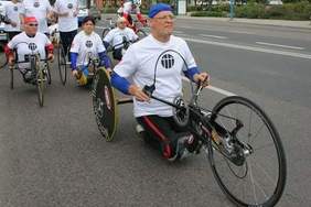 Pokaż zdjęcie: Alt: Zawody kolarskie. Na pierwszym planie bohater artykułu klęczy na stopniach roweru ręcznie napędzanego.  Na drugim planie trzy rowery ręczne prowadzone przez kobietę i mężczyzn. Na trzecim planie biegacze i kolarz na zwykłym rowerze szosowym.