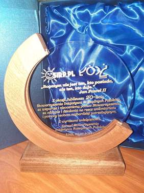 Pokaż zdjęcie: Na biurku stoi statuetka wręczana z okazji jubileuszu 20-lecia Stowarzyszenia Inicjatywa Rozsądnych Polaków. Za nią znajduje się opakowanie w granatowym kolorze. 