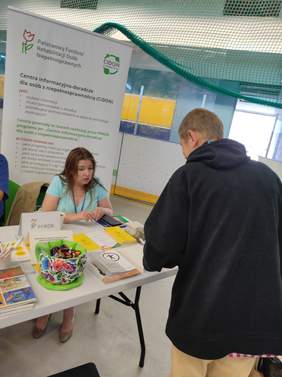 Pokaż zdjęcie: Przy stoisku informacyjnym PFRON pracownik udziela informacji zainteresowanej osobie