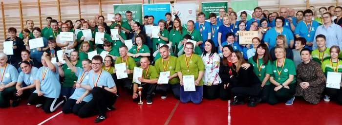 Pokaż zdjęcie: W sali gimnastycznej znajduje się grupa kilkudziesięciu osób ubrana w kolorowe stroje przyporządkowane do poszczególnych drużyn. Uczestnicy zawodów mają zawieszone na szyi medale. Część z tych osób trzyma w dłoniach także dyplomy.