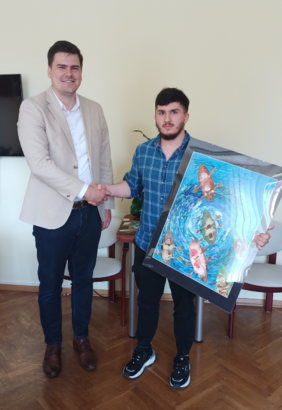 Pokaż zdjęcie: Dyrektor Oddziału ściska dłoń studentowi, który trzyma w drugiej ręce obraz z kajakarzami na wodzie widzianymi z góry