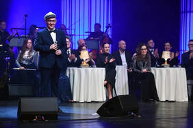 Mężczyzna w granatowym garniturze, trzyma w ręku mikrofon, na głowie ma białą czapkę kapitana, stoi na scenie