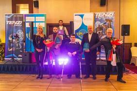 Pokaż zdjęcie: Grupa pięciu osób stoi na scenie podczas uroczystości wręczenia nagród. W centrum znajduje się mężczyzna korzystający z kul, który trzyma statuetkę. Po jego lewej stronie stoją kobieta w czarnym stroju z bukietem kwiatów oraz młody mężczyzna w marynarce trzymający nagrodę i kwiaty. Po prawej stronie znajdują się dwaj mężczyźni, jeden z nich (starszy, w okularach) trzyma bukiety kwiatów oraz torby upominkowe. W tle znajdują się banery z logotypami "TENZI" oraz „Ministerstwo Sportu i Turystyki”, sugerujące, że wydarzenie dotyczy sportu lub integracji. Na scenie widoczny jest również DJ lub prowadzący przy stanowisku z laptopem.
