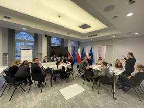 Pokaż zdjęcie: grupa około 15 osób uczestniczy w warsztatach w jasnym pomieszczeniu z flagami Polski i UE w tle. Uczestnicy siedzą przy stołach, jedna osoba gestykuluje, prowadząc dyskusję. Na podłodze widoczny jest arkusz z notatkami