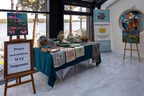 Pokaż zdjęcie: Wystawa prac uczestników WTZ w Augustowie. Po lewej stronie stoi sztaluga z obrazem przedstawiającym kwiaty, a poniżej znajduje się tablica z napisem „Prace uczestników WTZ Augustów”. Pośrodku na stole przykrytym zielonym obrusem wystawione są różne rękodzieła, takie jak ceramika, hafty oraz tkaniny. W tle, za dużym oknem, widać jezioro i drzewa. Po prawej stronie znajdują się dwie sztalugi z kolorowymi obrazami w stylu abstrakcyjnym. W tle widoczny jest baner z logo oraz nazwą WTZ Augustów.