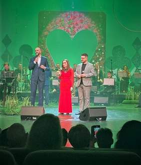 Artystka w czerwonej sukni stoi na śroku sceny, a po jej bokach dwóch panów - prowadzący koncert podczas występu na scenie