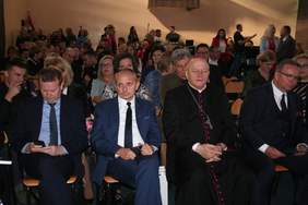 Pokaż zdjęcie: W sali gimnastycznej na krzesłach siedzą zaproszeni goście, bardzo elegancko ubrani. Na pierwszym planie biskup oraz zastępca prezydenta Lublina