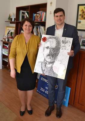 Pokaż zdjęcie: Na zdjęciu kobieta w średnik wieku i młody mężczyzna, który trzyma obraz. Uśmiechają się do obiektywu