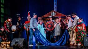 Pokaż zdjęcie: na zdjęciu widzimy scenę na której stoją osoby grupa wokalna, poprzebierani w kolorowe stroje, jedna z nich trzyma mikrofon i śpiewa do widowni. Tło jest w kolorze niebieskim 