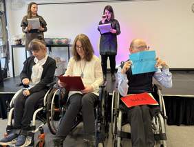 Pokaż zdjęcie: Na scenie dwie osoby prowadzą wystąpienie. Przed nimi siedzą trzy osoby na wózkach. Jedna trzyma kartkę z napisem „ZROZUMIENIE”. W tle widoczne stoisko z upominkami. Wydarzenie ma oficjalny i integracyjny charakter.