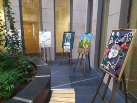 Pokaż zdjęcie: Na tle kamiennej ściany stoją sztalugi, a na nich są wyeksponowane obrazy, po lewej stronie zdjęcia kompozycja z roślin zielonych