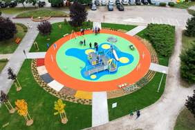 Pokaż zdjęcie: Z lotu ptaka widoczny jest plac zabaw w kształcie okręgu, który otoczony jest różnorodną roślinnością. Na placu zabaw znajduje się kilka osób. Do placu prowadzi pięć ścieżek. W tle można dostrzec parking wraz z samochodami osobowymi.