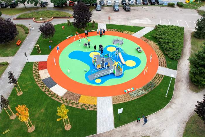 Pokaż zdjęcie: Z lotu ptaka widoczny jest plac zabaw w kształcie okręgu, który otoczony jest różnorodną roślinnością. Na placu zabaw znajduje się kilka osób. Do placu prowadzi pięć ścieżek. W tle można dostrzec parking wraz z samochodami osobowymi.