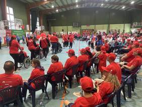 Pokaż zdjęcie: Na halę sportową, w której znajduje się grupa kilkudziesięciu osób, wchodzą sportowcy ubrani w jednolite stroje. Na obrzeżach hali ustawiona jest duża ilość banerów informujących o wydarzeniu oraz o podmiotach finansujących przedsięwzięcie.