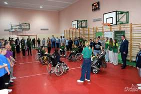 Pokaż zdjęcie: Grupa kilkudziesięciu osób stoi w hali sportowej. Po środku boiska znajduje się sześć osób w zielonych koszulkach, które poruszają się na wózku.