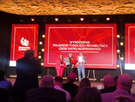 Uroczystość na scenie konferencyjnej z czerwonymi ekranami LED. Trzy osoby stoją na środku sceny i prezentują dyplomy oraz kwiaty. Na ekranie widoczny napis: „Wyróżnienie Polskiego Funduszu Rehabilitacji Osób Niepełnosprawnych”. Publiczność na pierwszym planie.