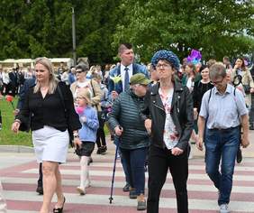 Pokaż zdjęcie: Po przejściu dla pieszych idzie tłum ludzi. Dużo jest osób z niepełnosprawnościami. Niektórzy trzymają w dłoniach kwiaty. 