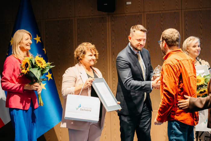 Pokaż zdjęcie: Cztery osoby stoją na scenie podczas uroczystości. Mężczyzna w ciemnym garniturze (prawdopodobnie ten sam co na zdjęciu 2) podaje, lub odbiera, nagrodę (małą, przezroczystą statuetkę) mężczyźnie ubranemu w jasnopomarańczową, sportową bluzę. Po lewej stronie stoi uśmiechnięta kobieta w jasnoróżowej marynarce, która trzyma szarą torbę z logo "Pomorze Zachodnie" oraz szarą ramkę z dyplomem. Obok niej, po jej lewej stronie, jest uśmiechnięta kobieta w jaskrawo różowej marynarce i niebieskich spodniach, trzymająca duży bukiet słoneczników. Za nią widać fragment flagi Unii Europejskiej. Po prawej stronie, obok mężczyzny w pomarańczowej bluzie, widoczna jest kolejna uśmiechnięta kobieta.
