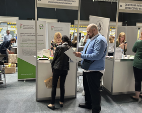 Pokaż zdjęcie: Przed stoiskiem z napisem Państwowy Fundusz Rehabilitacji Osób niepełnosprawnych stoją dwie osoby, za nim stoi kobieta w sukience w kwiaty. Na stole leża materiały prasowe z logo PFRON. Za stoiskiem stoją dwa banery z logo PFRON.