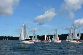 Pokaż zdjęcie: Na jeziorze na pierwszym planie płynie 6 jechatów, na drugim planie o 4 jachtów. Niebieskie niebo z białymi chmurami