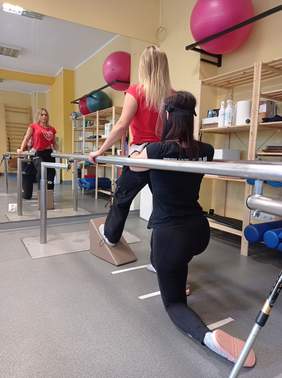 Pokaż zdjęcie: fragment sali rehabilitacyjnej przed dużym lustrem ćwiczy kobieta na stojąco druga ja podtrzymuje