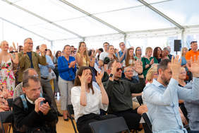 Pokaż zdjęcie: Duży biały namiot, kilkadziesiąt osób siedzi lub stoi klaszcząc lub machając uniesionymi do góry dłoniami