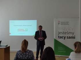 Pokaż zdjęcie: Mężczyzna w garniturze stoi przez osobami siedzącymi na krzesłach po prawej stronie stoi baner z logo PFRON i napisem wszyscy jesteśmy tacy sami w tle na ścianie wyświetlony jest tytułowy slajd prezentacji