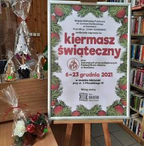 Pokaż zdjęcie: Fragment pomieszczenia na pierwszym planie duży plakat z zaproszeniem na świąteczny kiermasz w bibliotece filia nr 20 w Sosnowcu do 23 grudnia. Plakat umieszczony jest na sztaludze, po lewej stronie na stole stoją zapakowane ozdoby świąteczne.