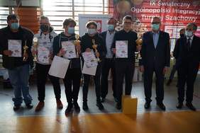 Pokaż zdjęcie: zdjęcie grupowe na tle okien w sali gimnastycznej stoją zwycięzcy turnieju 3 młodych chłopców i 2 mężczyzn trzymają w rękach nagrody obok stoi 3 mężczyzn w garniturach 