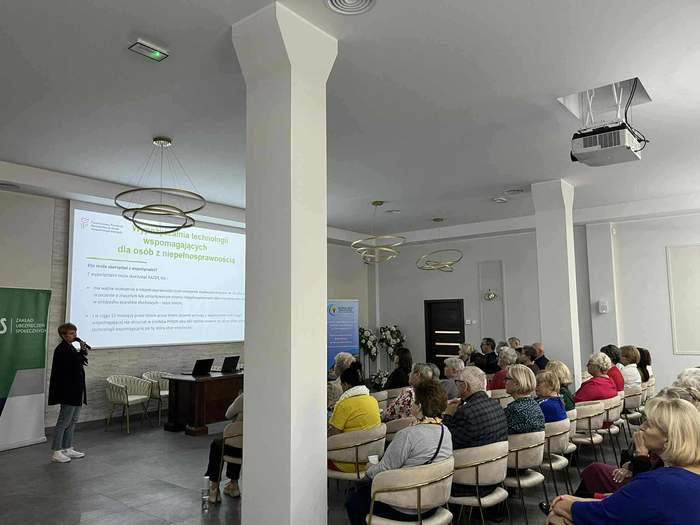 Grupa osób, siedzą, jedna kobieta stoi, w tle prezentacja