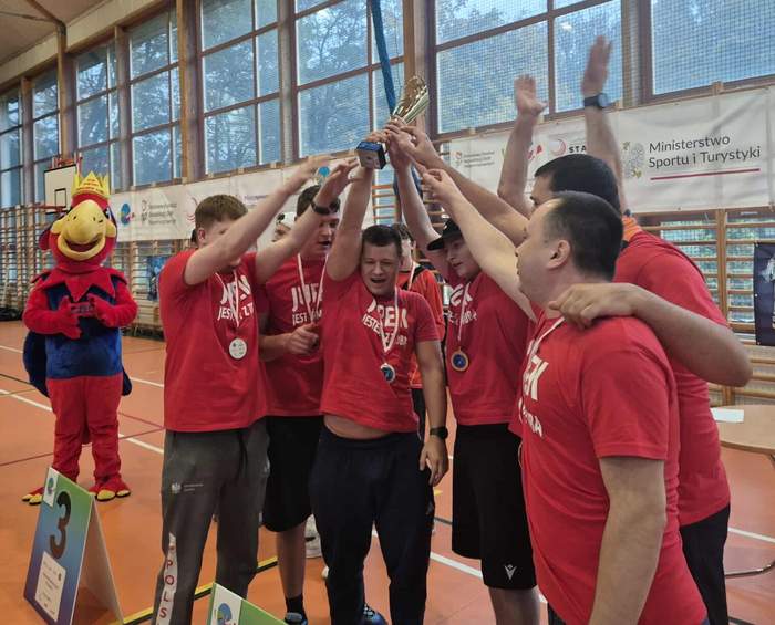 Drużyna w czerwonych koszulkach z medalami na szyjach, wznosząca wspólnie duży złoty puchar w hali sportowej. Wszyscy są uśmiechnięci i radośnie celebrują. Po lewej stronie stoi maskotka w przebraniu czerwono-niebieskiego ptaka. W tle widać siatkę okienną i banery.