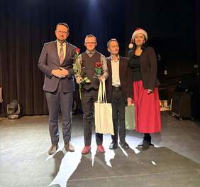 Pokaż zdjęcie: Czworo osób stoi w rzędzie na scenie podczas oficjalnej części wydarzenia. Od lewej: mężczyzna w ciemnym garniturze i okularach, mężczyzna w kamizelce trzymający czerwoną różę oraz statuetkę w kształcie tulipana, mężczyzna w brązowej marynarce trzymający białą torbę oraz kobieta w czerwonej spódnicy i czapce świętego Mikołaja. Wszyscy patrzą w stronę obiektywu. Scena jest ciemna, oświetlona jedynie od góry reflektorami.