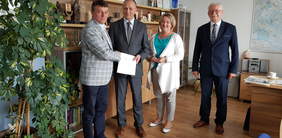 Cztery osoby, trzech mężczyzn i jedna kobieta, stoją na tle regału, uśmiechają się, dwóch mężczyzn przekazuje sobie dokument i ściska dłonie, w lewym rogu zdjęcia wysoki zielony kwiat, z prawej fragment biurka