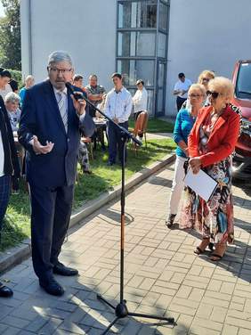 Pokaż zdjęcie: Przy mikrofonie stoi mężczyzna, który wygłasza przemówienie. Wokół niego stoją ludzie. 