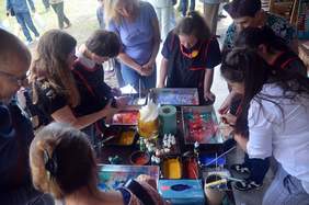 Pokaż zdjęcie: Przy stole grupa młodzieży maluje farbami na blachach do pieczenia