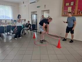 Pokaż zdjęcie: Zawody z użyciem hula-hop w jasnej sali. Na środku stoi pochylony mężczyzna z dwoma różowymi kołami hula-hop – jednym wokół bioder, drugim w dłoniach. Przy jego stopach stoją dwa pomarańczowe pachołki wyznaczające tor. Po prawej stoi drugi mężczyzna w szarej koszulce i ciemnych szortach, który mierzy czas telefonem. W tle po lewej grupa osób przy stołach, w tym osoba na wózku.