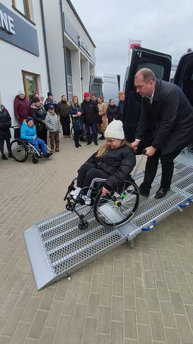 Pokaż zdjęcie: Mężczyzna pomaga dziewczynce na wózku inwalidzkim zjechać po metalowej rampie z samochodu dostawczego. W tle stoi grupa ludzi, obserwują wydarzenie przed budynkiem.