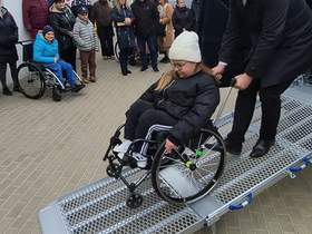 Mężczyzna pomaga dziewczynce na wózku inwalidzkim zjechać po metalowej rampie z samochodu dostawczego. W tle stoi grupa ludzi, obserwują wydarzenie przed budynkiem.