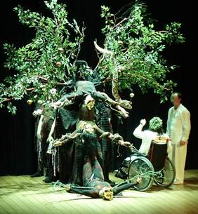 Pokaż zdjęcie: Na scenie trwa występ artystyczny. Na środku stoi drzewo, a wokół niego znajdują się aktorzy. Na gałęziach wiszą czerwone jabłka oraz wąż. 