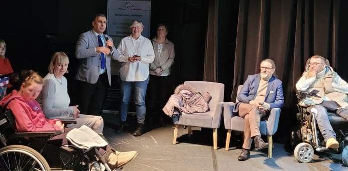 Pokaż zdjęcie: Mężczyzna w marynarce trzyma mikrofon. Po prawej jego stronie stoi kobieta i trzy fotele, na których siedzi mężczyzna i kobieta. Po lewej stronie osoby na wózkach inwalidzkich.