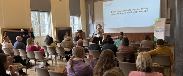 Pokaż zdjęcie: Sala z wysokimi oknami, uczestnicy siedzą na ustawionych w rzędach krzesłach. Przed nimi stoi prelegent w jasnej marynarce, mówiący do mikrofonu. W tle duży ekran z prezentacją o dofinansowaniu wynagrodzeń pracowników z niepełnosprawnością oraz baner PFRON.