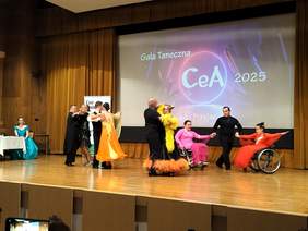 Pokaż zdjęcie: Scena pełna tańczących osób. Tancerze są ubrani w piękne barwne kreacje. Wśród nich są osoby z zespołem Downa oraz na wózku inwalidzkim. 