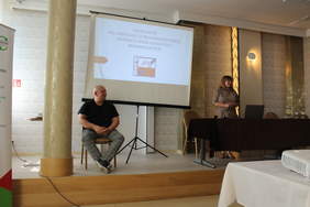 Pokaż zdjęcie: W sali konferencyjnym po lewej stronie siedzi na krześle tłumacz języka migowego, po prawej za stołem prezydialnym siedzi kobieta z mikrofonem, w tle po lewej na ekranie wyświetlana jest prezentacja