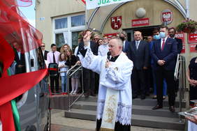 Pokaż zdjęcie: Ksiądz trzyma kropidło,którym święci nowe auta .W tle budynek szkoły  przed którym stoją zgromadzeni goście oraz dzici ze szkoły