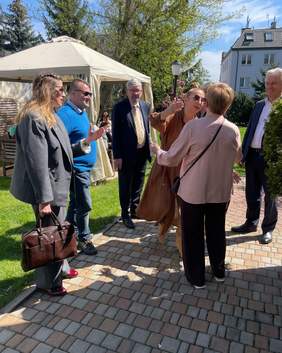 Pokaż zdjęcie: W ogrodzie stoją elegancko ubrani ludzie. W tle widać wielkanocne dekoracje. Niektórzy witają się ze sobą. 