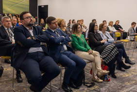 Pokaż zdjęcie: Widok sali konferencyjnej. Kobiety i mężczyźni siedzą na krzesłach ustawionych w rzędach. Patrzą przed siebie. Słuchają prezentacji. Uśmiechają się.