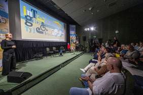 Pokaż zdjęcie: Osoba prowadząca wydarzenie stoi na scenie obok dużego ekranu z kolorową grafiką festiwalu. Na ekranie widnieje napis „22 Europejski Festiwal Filmowy Integracja Ty i Ja, Koszalin 2–6.09.2025” oraz ilustracja plaży z parasolem i leżakami, z których jeden ma koła jak wózek inwalidzki. Publiczność siedzi w sali kinowej i bije brawo.