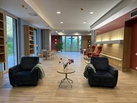 Pokaż zdjęcie: Duże pomieszczenie, na podłodze kafle we wzorze drewna, na ścianach drewniane półki i drewniane wykończenia. Na środku dwa fotele na których leża koce, miedzy nimi stolik na którym stoją kwiaty. Po lewej stronie i na końcu pomieszczenia duże 