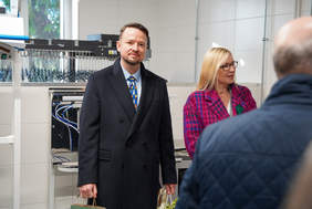 Dwóch ludzi stoi w jasnym, czystym pomieszczeniu, prawdopodobnie pralni lub zakładzie produkcyjnym. Mężczyzna w granatowym płaszczu i krawacie patrzy wprost, a kobieta w różowo-fioletowym płaszczu w kratę spogląda w prawo, częściowo zasłonięta przez niewyraźną postać. W tle widoczne jest profesjonalne wyposażenie.