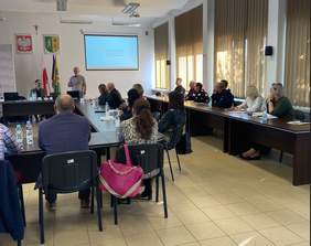 Pokaż zdjęcie: Sala konferencyjna, stoły ustawione w podkowę. Na ekranie wyświetlana jest prezentacja, przy stole siedzą słuchacze.